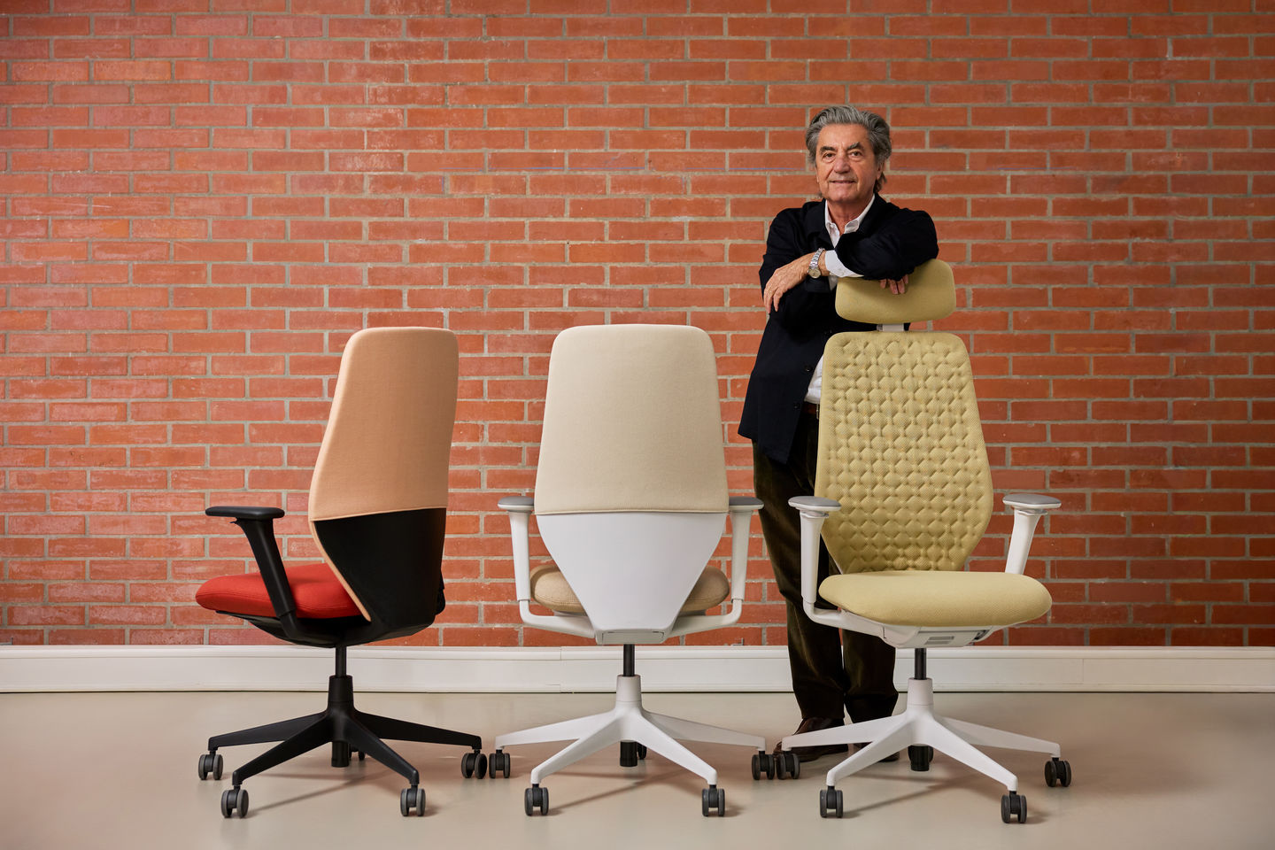 Drei Vitra Bürostühle in unterschiedlichen Ausführungen stehen vor einer Backsteinwand, daneben lehnt eine Person an einem Modell mit gesteppter Rückenlehne.